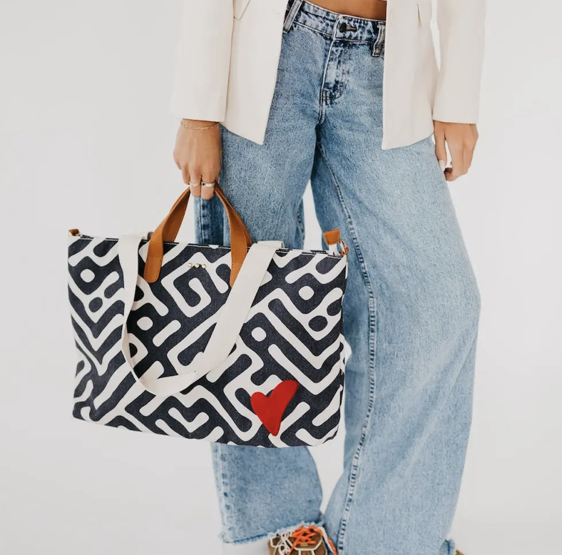 A person holding a black and white geometric patterned canvas tote bag with tan handles and a red heart accent.