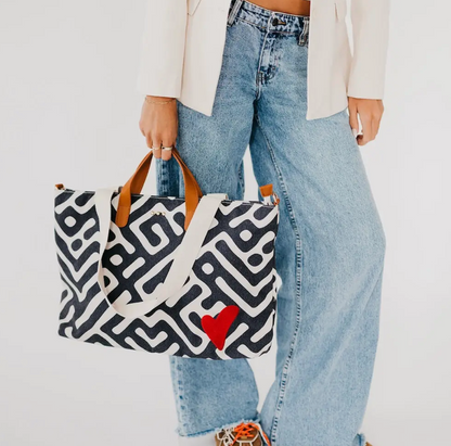 A person holding a black and white geometric patterned canvas tote bag with tan handles and a red heart accent.