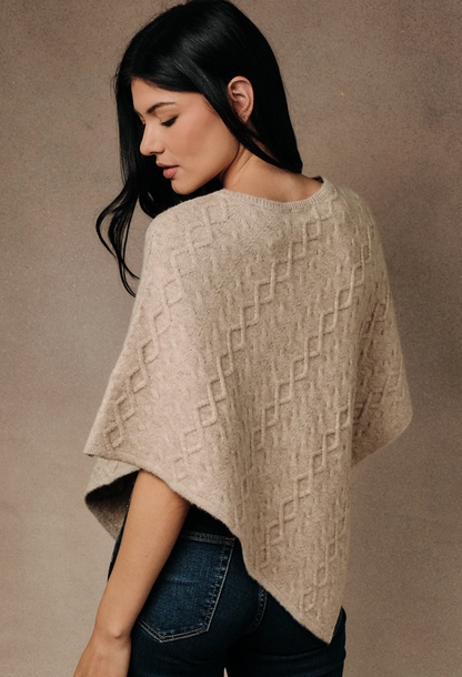 A woman wearing a beige textured knit poncho sweater and dark blue jeans, photographed from a side-back angle against a neutral textured studio background.