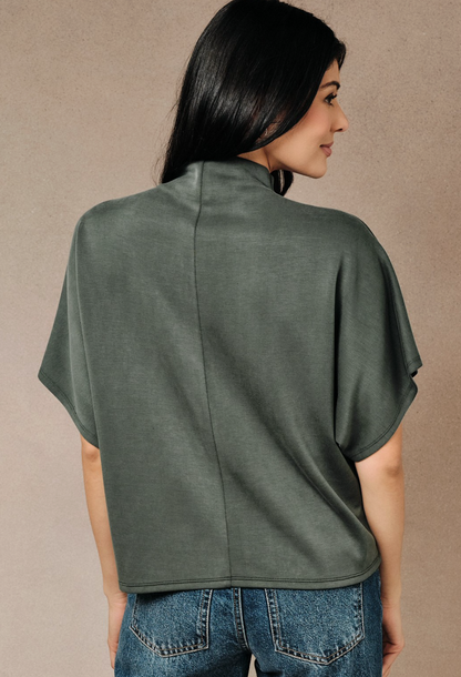 A woman shown from the back wearing a Olive dolman short sleeve top with lightweight drape and relaxed silhouette., paired with blue jeans, against a neutral textured beige background.