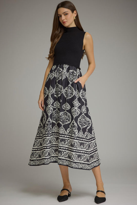 A woman modeling a sleeveless midi dress with a solid black mock neck bodice and a black and white geometric patterned skirt, paired with black flats against a neutral gray studio background.