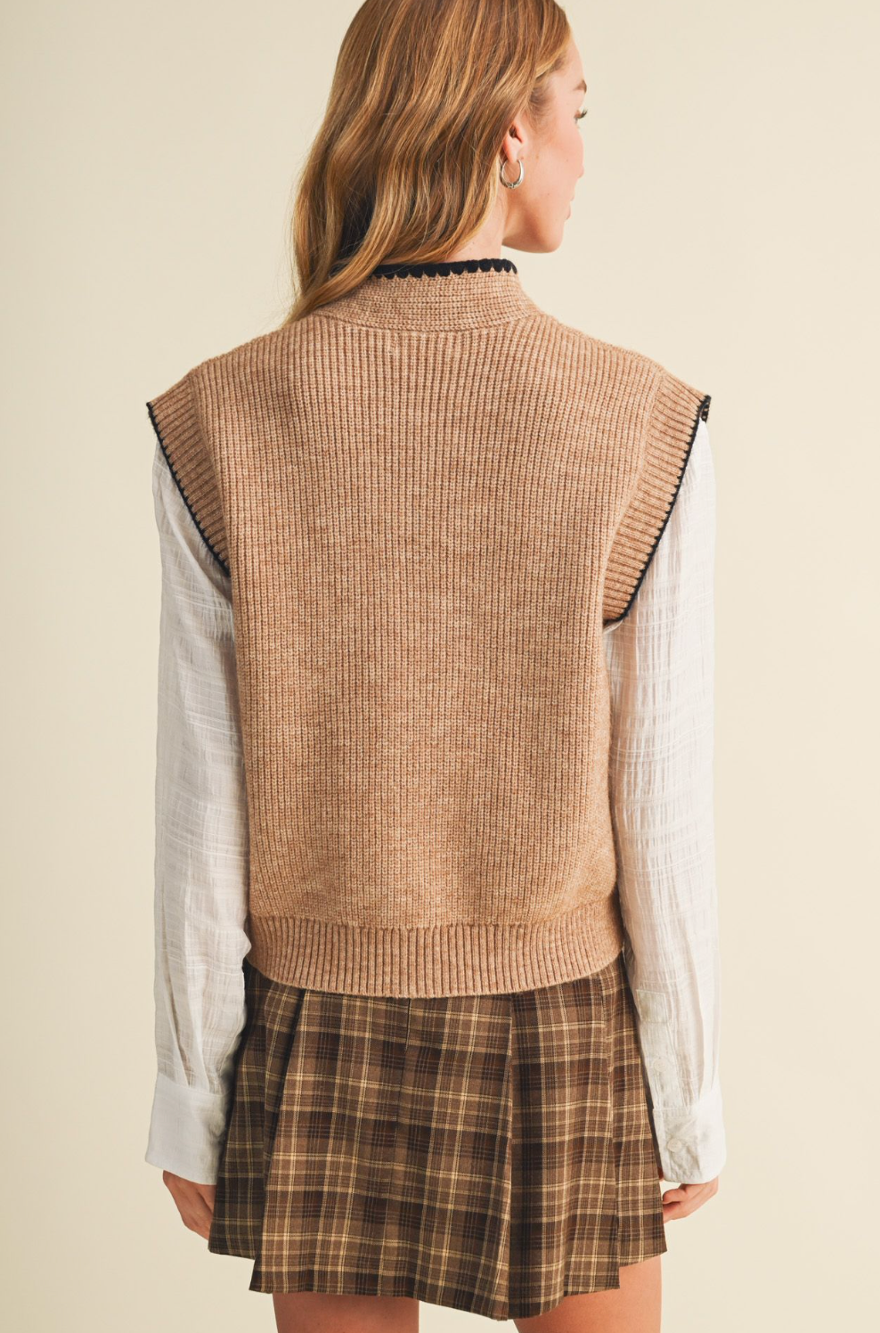 A woman shown from the back wearing a Mustard Seed sleeveless ribbed knit sweater vest in a warm tan/beige color, layered over a white long-sleeve shirt and a brown plaid pleated skirt.