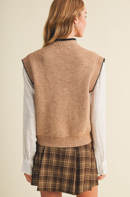 A woman shown from the back wearing a Mustard Seed sleeveless ribbed knit sweater vest in a warm tan/beige color, layered over a white long-sleeve shirt and a brown plaid pleated skirt.