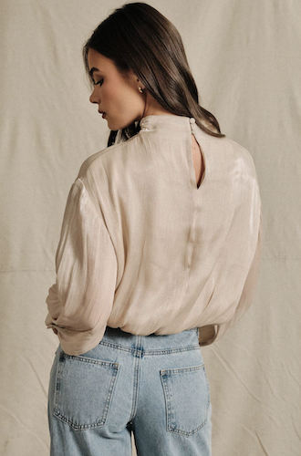 A woman shown from the back wearing a light beige high-neck blouse with a small keyhole cutout at the back, paired with light blue jeans, against a neutral draped fabric backdrop.