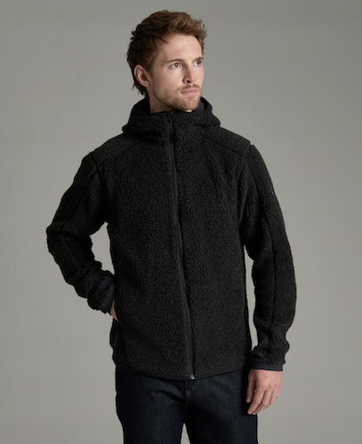 A man wearing a black Kuhl Skapegoat textured fleece zip-up hoodie jacket, paired with dark blue jeans, standing against a solid gray studio background.