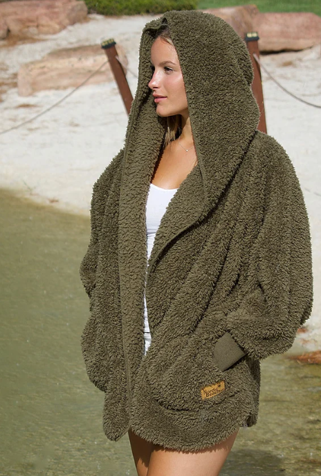 A woman wearing a plush, textured Nordic Beach olive green hooded wrap standing outdoors near shallow water and a sandy dock area.