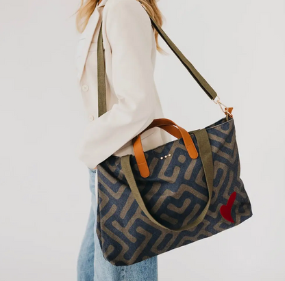 A person carrying a large patterned canvas tote bag with olive green handles and strap, featuring a small red heart detail on the side, against a plain white background.