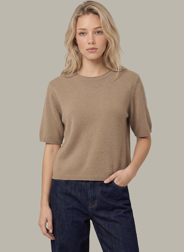 A woman modeling a short-sleeve crewneck knit sweater in a warm taupe-brown color, paired with dark blue jeans, against a solid light gray background.