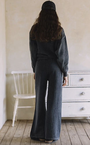 A person viewed from behind wearing a dark charcoal gray sweatshirt and matching wide-leg pants, standing in a room with a white chair and dresser.