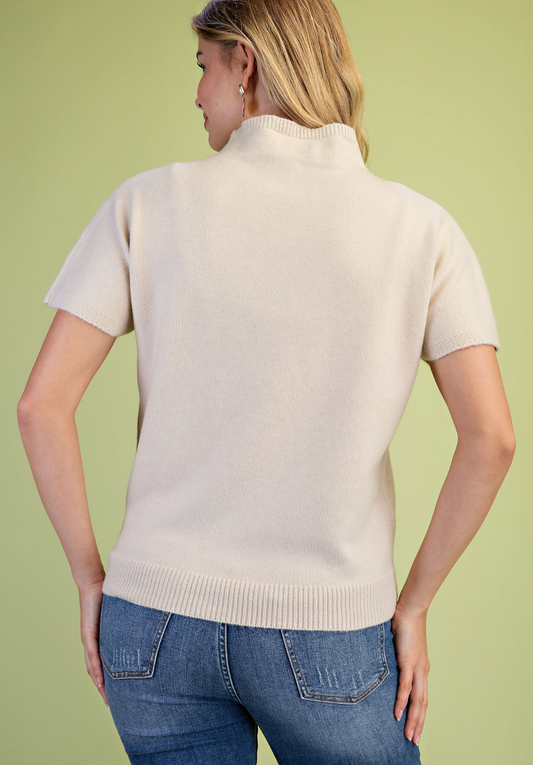 A woman shown from the back wearing a cream-colored short-sleeve mock neck knit sweater and blue jeans, standing against a solid light green background.