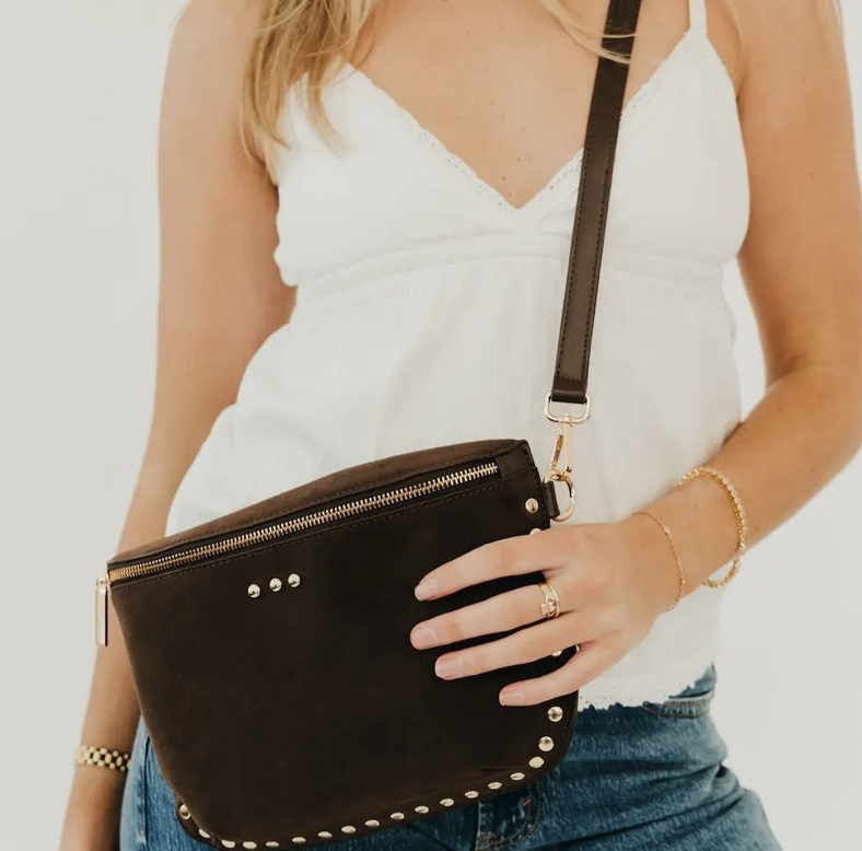 A woman wearing a white camisole and blue jeans carries a dark brown suede crossbody bag with gold-tone hardware and studded details.