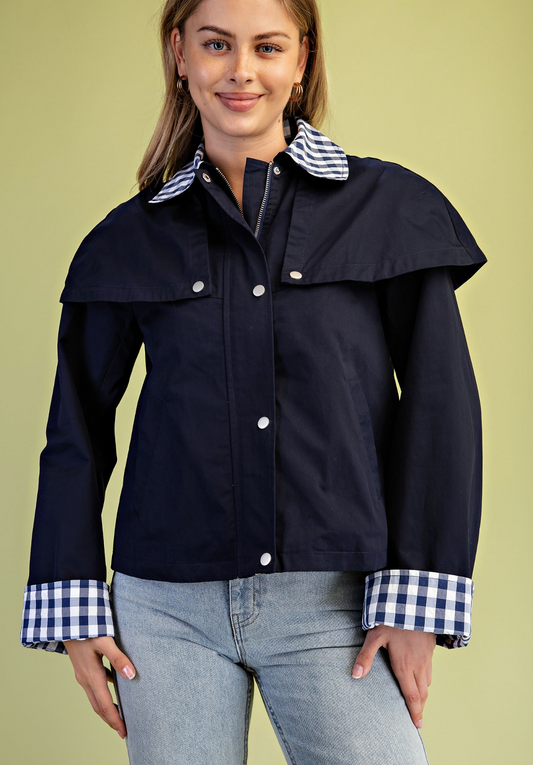 A woman wearing a Gigio navy blue jacket with a contrasting blue and white gingham check collar and cuffs, paired with light-wash jeans, against a solid pale yellow-green background.