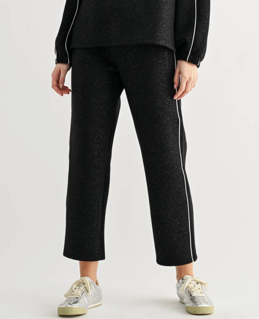 A model wearing glitter black cropped pants with white side stripes, paired with a matching black top and silver sneakers, shown from the waist down on a light gray background.