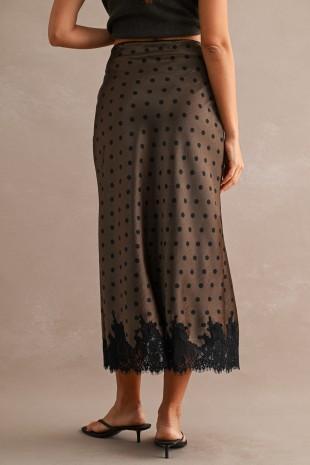 A woman wearing a sheer brown midi skirt with black polka dots and a black lace hem, paired with black flat sandals, shown from the back against a neutral beige background.