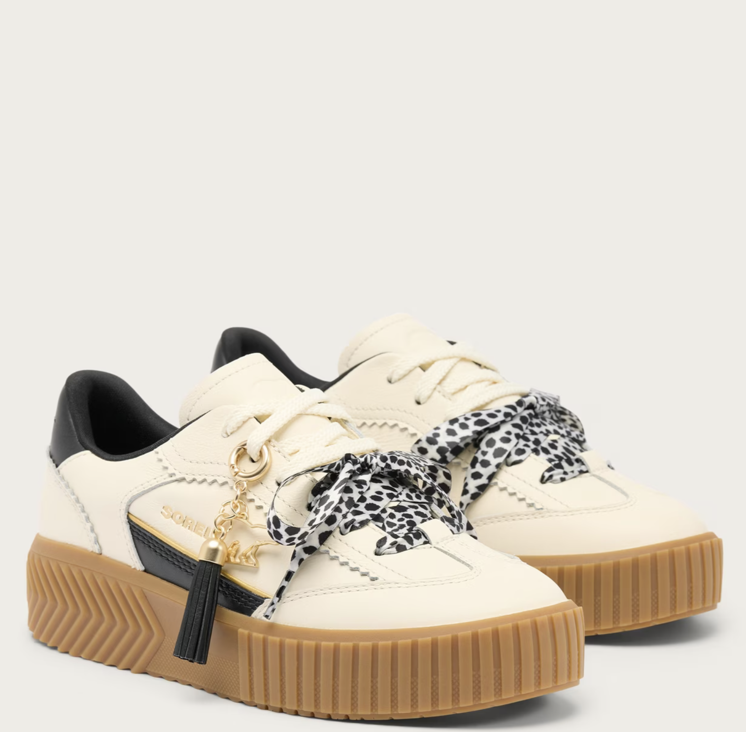 A pair of Sorel low-top platform sneakers in off-white leather with black leather heel tabs, brown gum rubber soles, and decorative black-and-white leopard-print ribbon bows on the laces. A gold-tone tassel charm with a black tassel and a gold brand name plate is attached to the side of the front shoe.