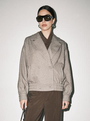 A model wearing a light brown double-breasted cropped jacket over a dark brown turtleneck, paired with dark brown trousers and black sunglasses, standing against a plain light gray background.