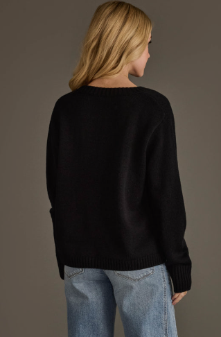 A woman shown from the back wearing a black knit sweater and light blue jeans against a plain taupe studio background.