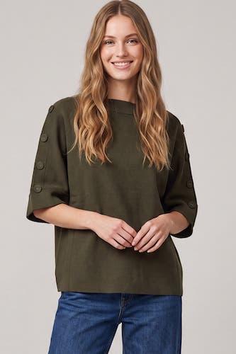 A woman with long wavy blonde hair wearing an olive green short-sleeve top with decorative buttons on the sleeves, paired with blue jeans, against a solid light gray background.