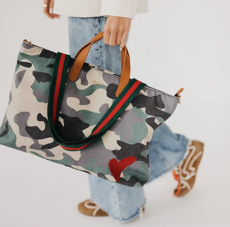 A person carrying a large camouflage tote bag with green and red web-style handles, light brown leather handles, and a red heart detail on the front, against a clean white background.