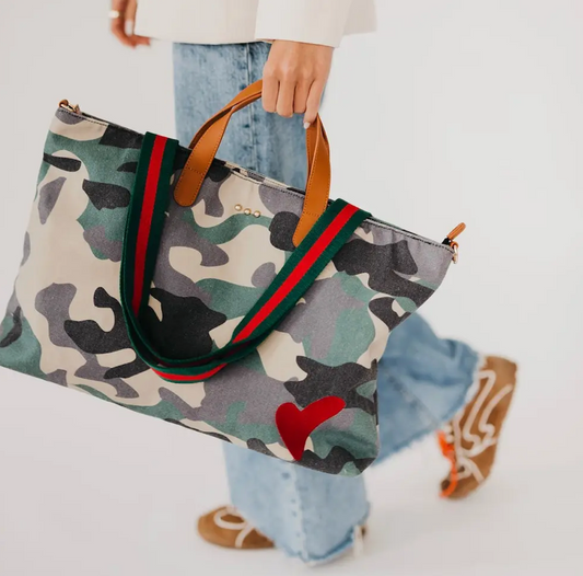 A person carrying a large camouflage tote bag with green and red web-style handles, light brown leather handles, and a red heart detail on the front, against a clean white background.