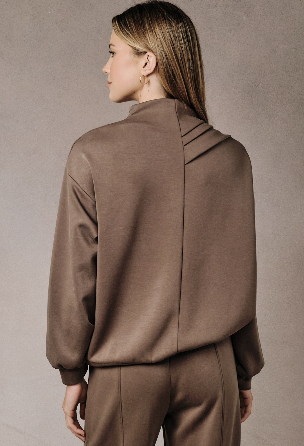 A woman shown from the back wearing a brown draped mock-neck top and matching brown trousers against a textured gray studio background.