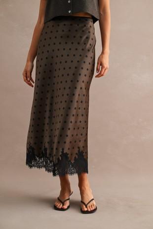 A model wearing a brown sheer midi skirt with black polka dots and a black lace hem, paired with black sandals against a textured beige studio background.