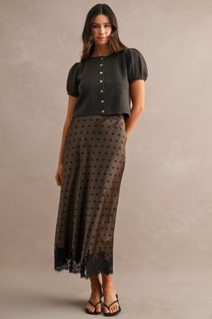 A woman modeling a dark gray short-sleeve button-front top paired with a brown sheer midi skirt with black polka dots and a black lace hem, against a textured beige studio background.