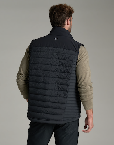 A man shown from the back wearing a Kuhl Spyfire black quilted puffer vest over a beige long-sleeve shirt and dark pants, photographed against a neutral gray studio background.