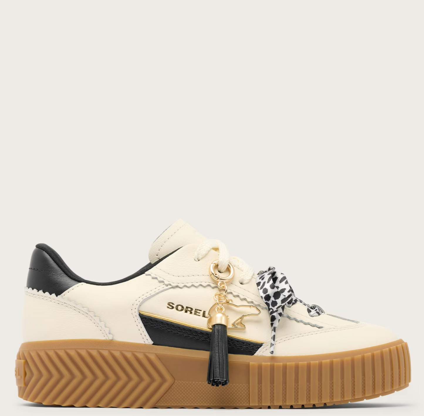 A Sorel low-top platform sneaker in off-white leather with black leather accents, a thick brown gum sole, and a decorative black-and-white patterned ribbon tied to the laces with a gold tassel charm. The brand name 'SOREL' is visible on the side.