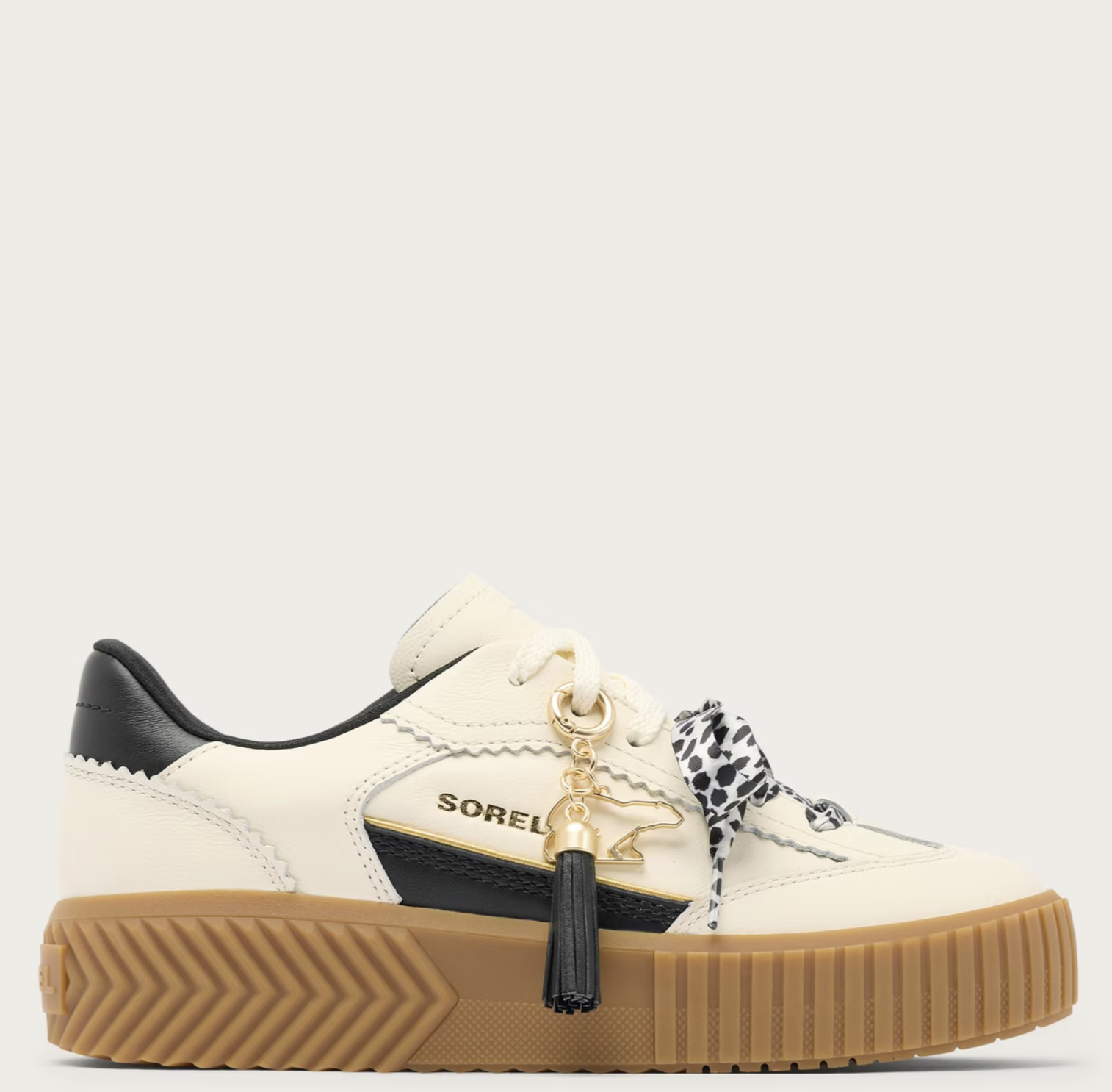 A Sorel low-top platform sneaker in off-white leather with black leather accents, a thick brown gum sole, and a decorative black-and-white patterned ribbon tied to the laces with a gold tassel charm. The brand name 'SOREL' is visible on the side.