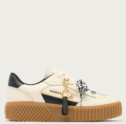 A Sorel low-top platform sneaker in off-white leather with black leather accents, a thick brown gum sole, and a decorative black-and-white patterned ribbon tied to the laces with a gold tassel charm. The brand name 'SOREL' is visible on the side.