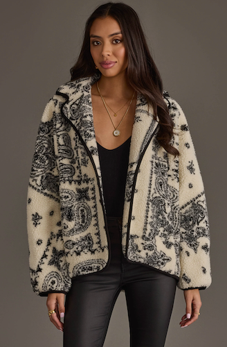 A woman modeling a cream-colored sherpa zip-up hoodie jacket with a black paisley pattern, worn over a black top and black pants against a solid dark gray studio background.