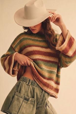 A woman wearing a chunky knit striped sweater with green, rust, and beige horizontal stripes, paired with an off-white wide-brim hat and olive green cargo shorts, set against a plain beige background.