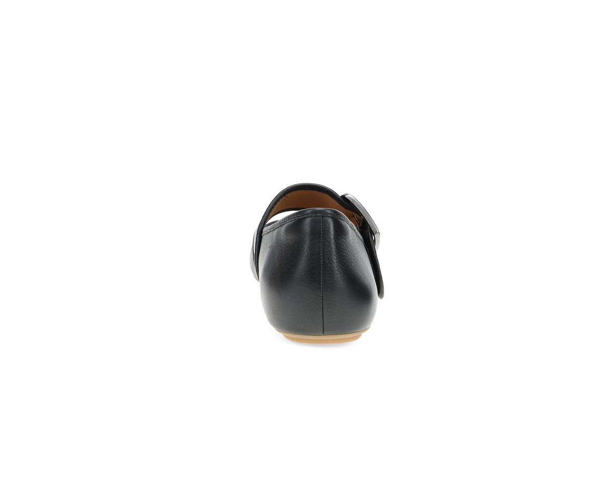 A single Dansko Matilda black leather flat shoe shown from the back, featuring a strap with a silver buckle and a light tan outsole, set against a plain white background.