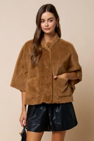 A woman modeling a cropped, short-sleeved brown faux fur jacket with front patch pockets, paired with black faux leather shorts, against a solid beige background.