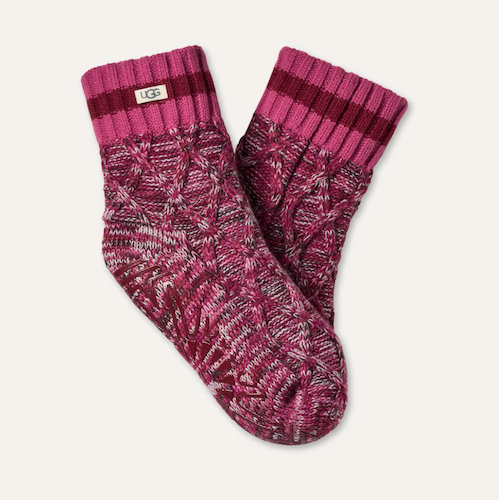 A pair of UGG cable-knit socks in a heathered magenta and burgundy colorway, featuring a ribbed cuff with a small white UGG logo tag, set against a light off-white background.