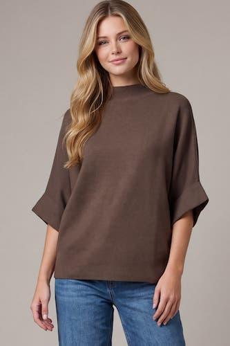 A woman modeling a brown short-sleeve knit top with a mock neck and wide elbow-length sleeves, paired with blue jeans, against a plain beige studio background.