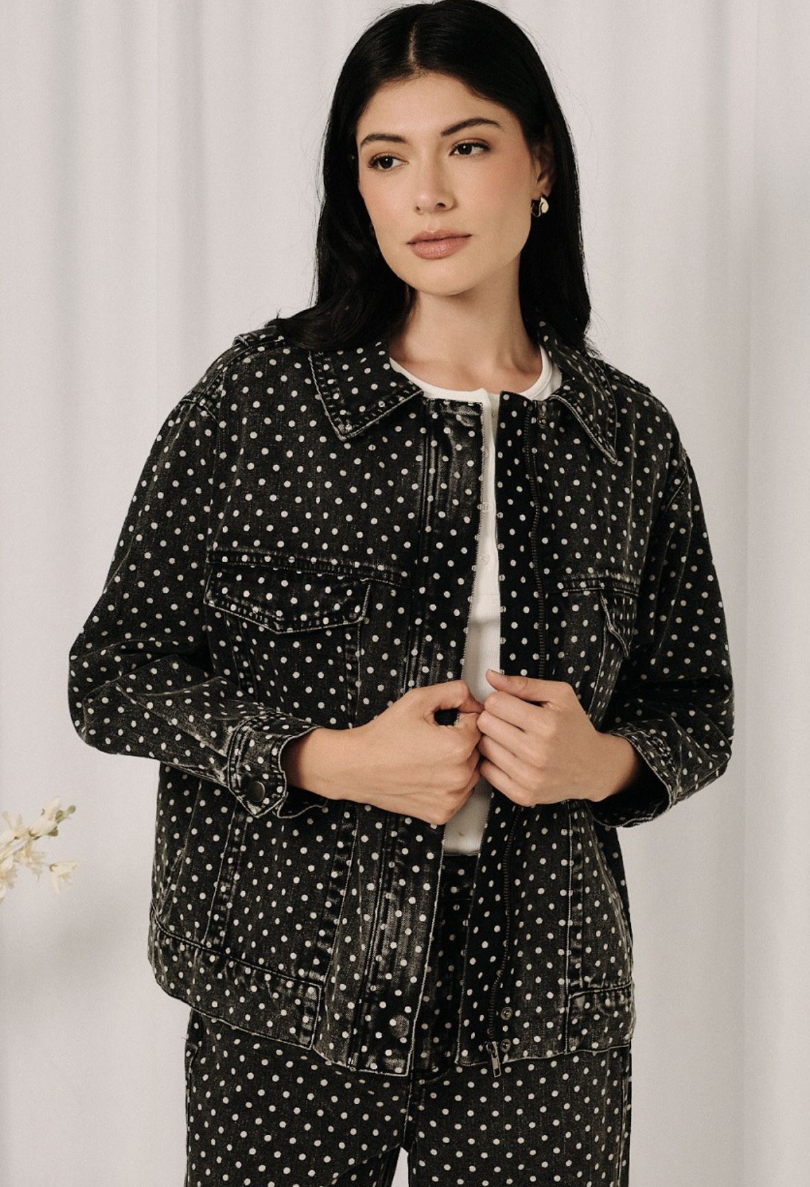 A woman wearing a black denim jacket and matching pants with a white polka dot pattern, styled over a white crewneck shirt, standing against a soft white curtain background.