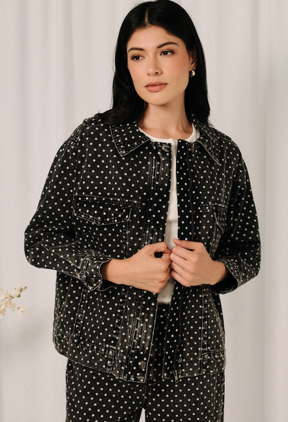 A woman wearing a black denim jacket and matching pants with a white polka dot pattern, styled over a white crewneck shirt, standing against a soft white curtain background.