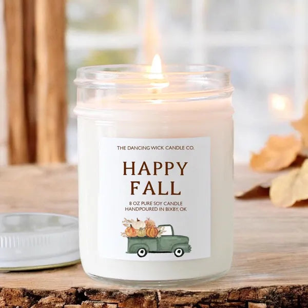 A lit 8 oz pure soy candle in a clear glass jar with a white label reading 'The Dancing Wick Candle Co. Happy Fall' and a small green truck carrying pumpkins, set on a rustic wood surface with blurred autumn leaves in the background.